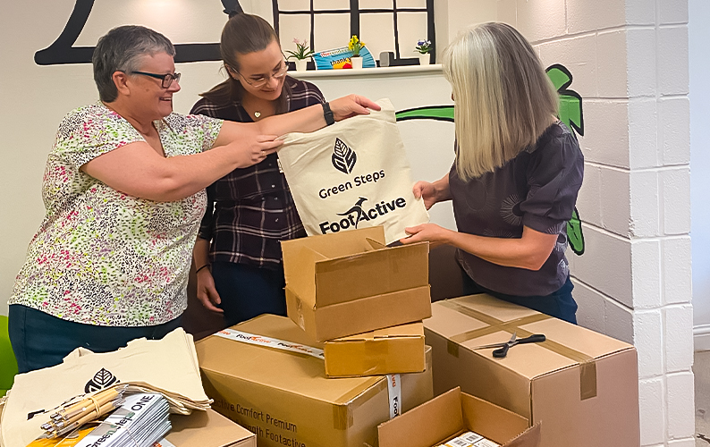 Our team members, Laura and Lynne handing Fiona our branded tote bag, surrounded by donation boxes.